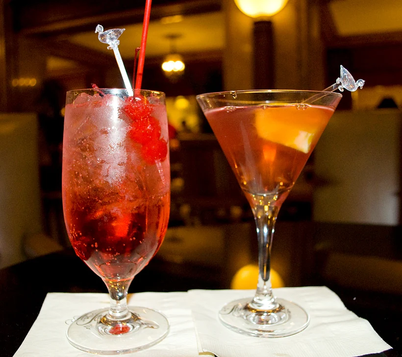 Ly Shirley Temple màu hồng đỏ bên cạnh một ly cocktail màu cam trên quầy bar