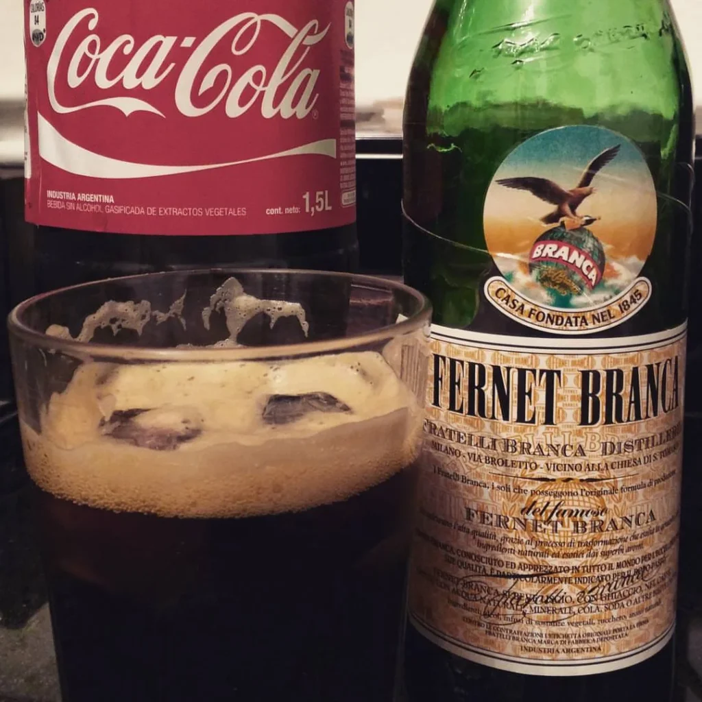 Ly Fernet pha Coca-Cola có đá, đặt cạnh chai Fernet-Branca và chai Coca-Cola